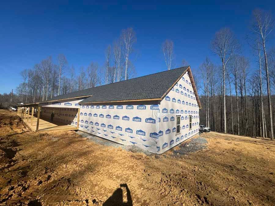 A house under construction wrapped in blue tarp with a dark roof on a hillside with bare trees.