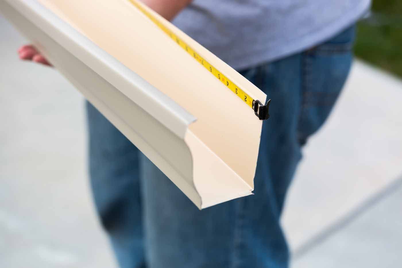 Person holding a section of light beige gutter, measuring with a tape.
