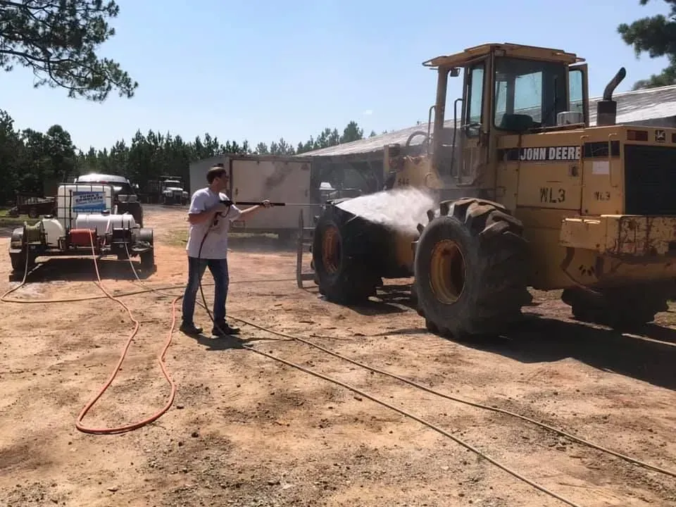 A person power washes a large yellow John Deere tractor outside on a sunny day.