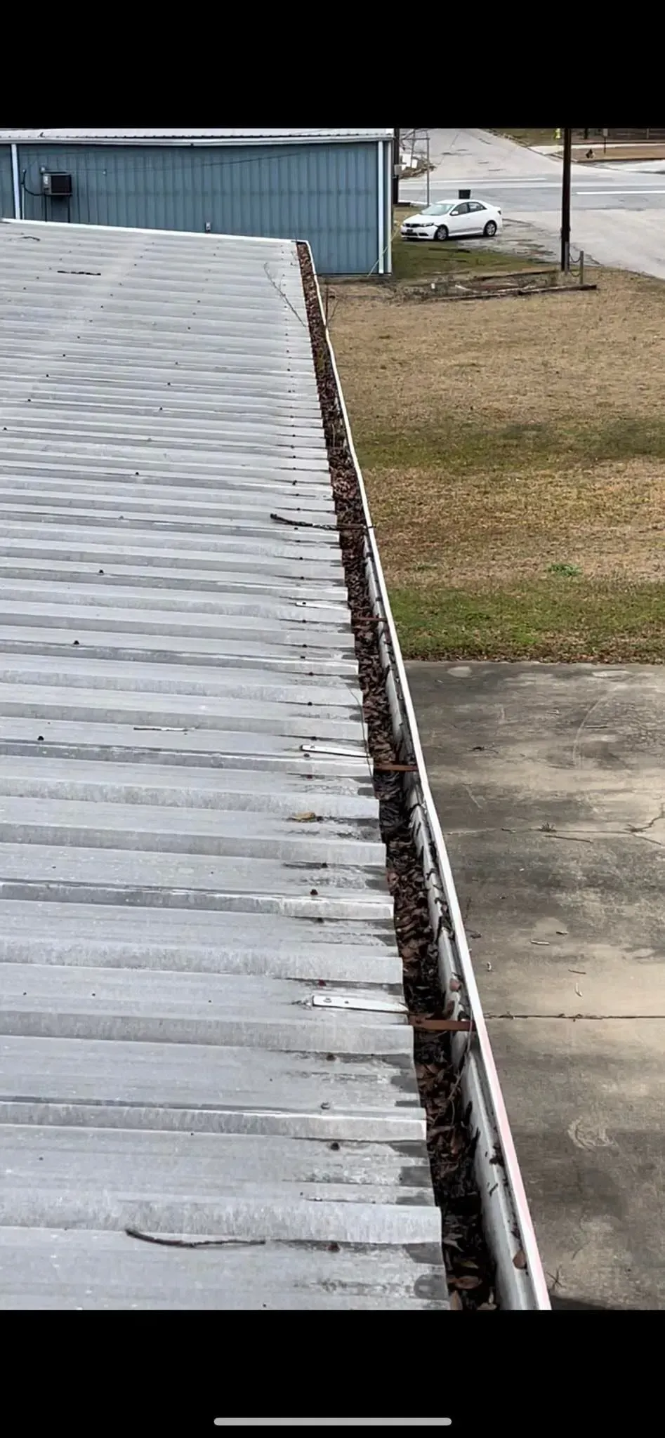 View of a roof with a gutter filled with debris. A vehicle sits on the adjacent grass.