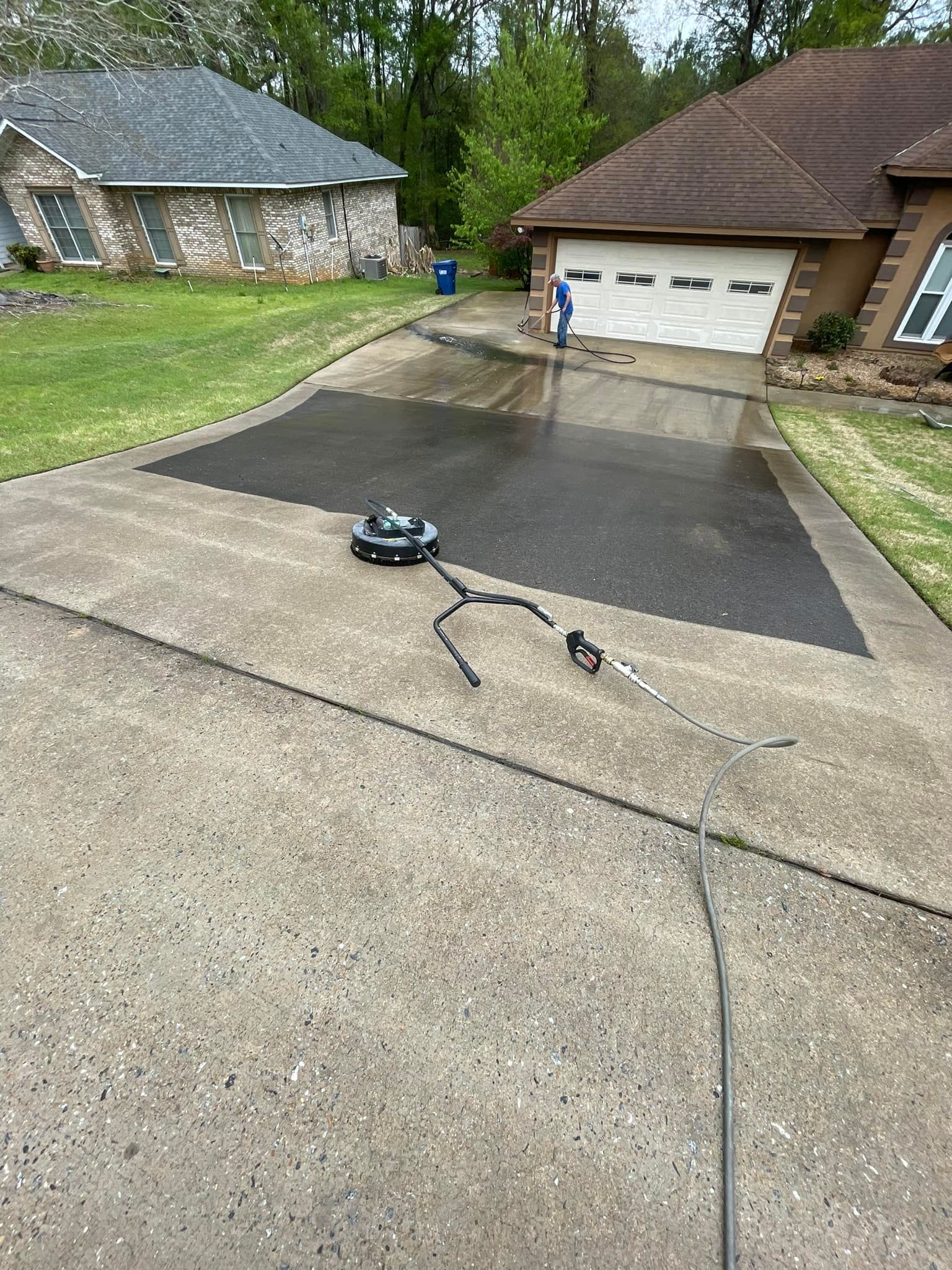 Person pressure washing a driveway with a surface cleaner; house and lawn in background.