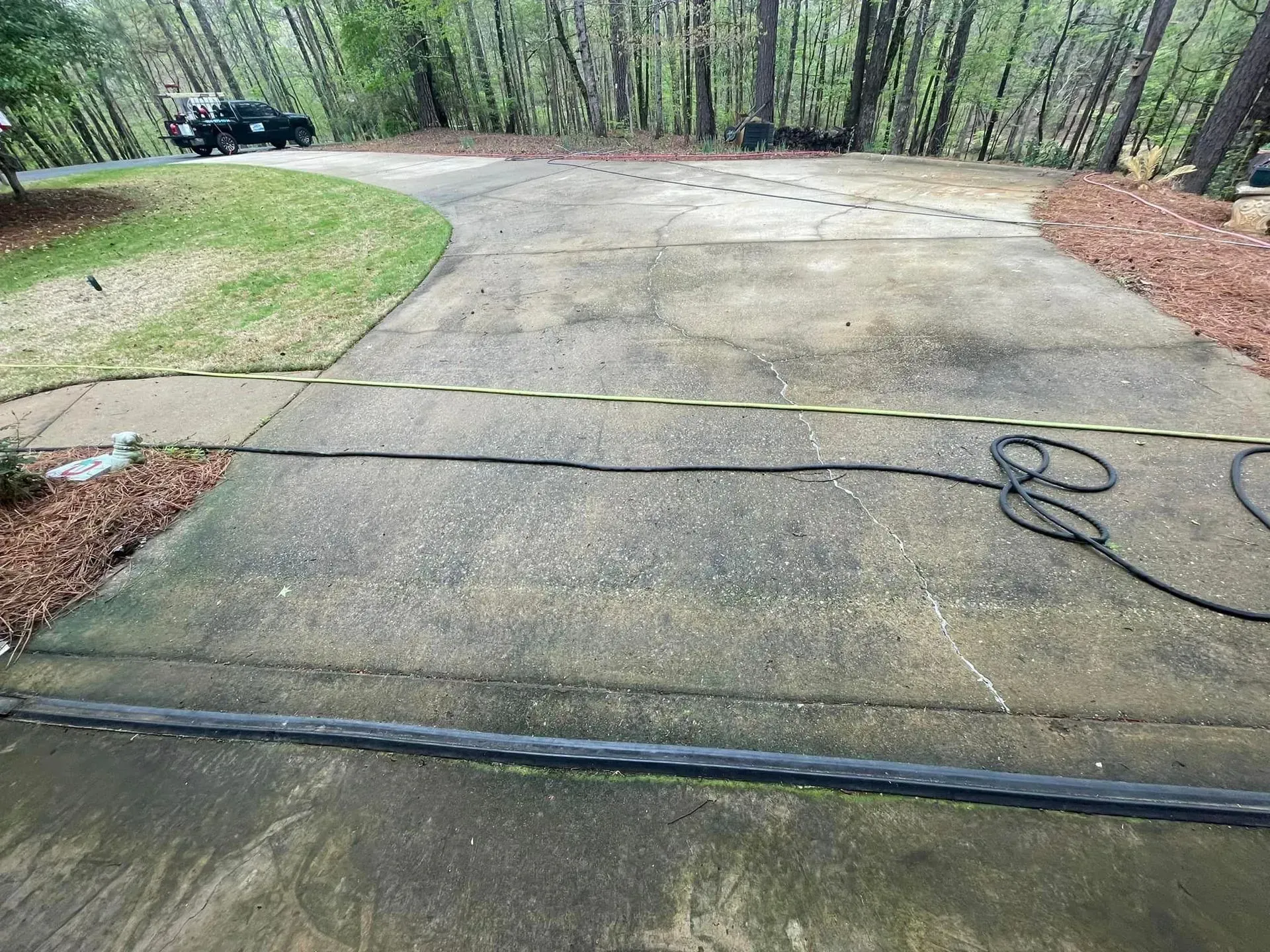 Driveway with a vehicle at the top, bordered by grass and trees. A hose lies on the concrete.