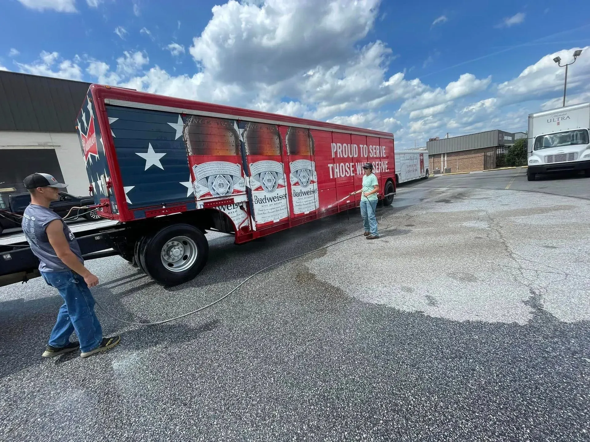 Two men push a red truck with patriotic beer can art on a gravel lot under a cloudy sky.