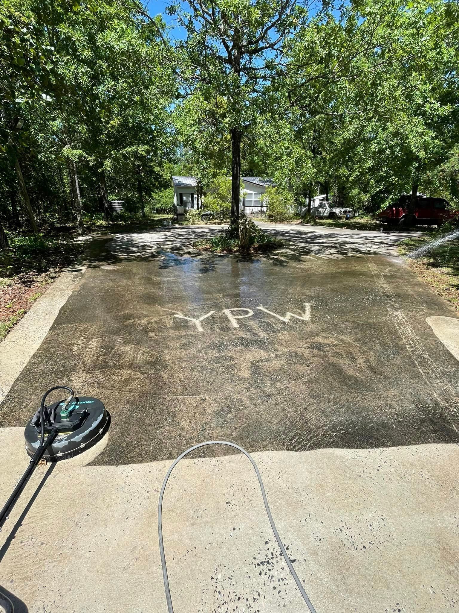 Pressure washing a concrete driveway; circular cleaner sprays water. Trees and house in background.