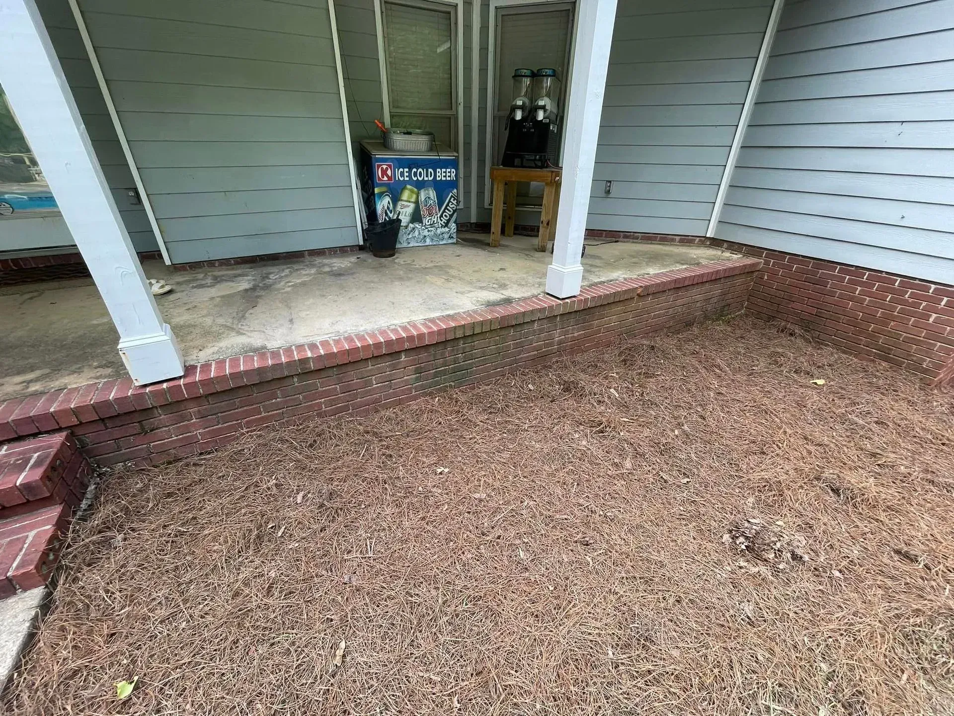 Exterior porch with brick border and brown mulch, a raised concrete patio, and a light blue house exterior.