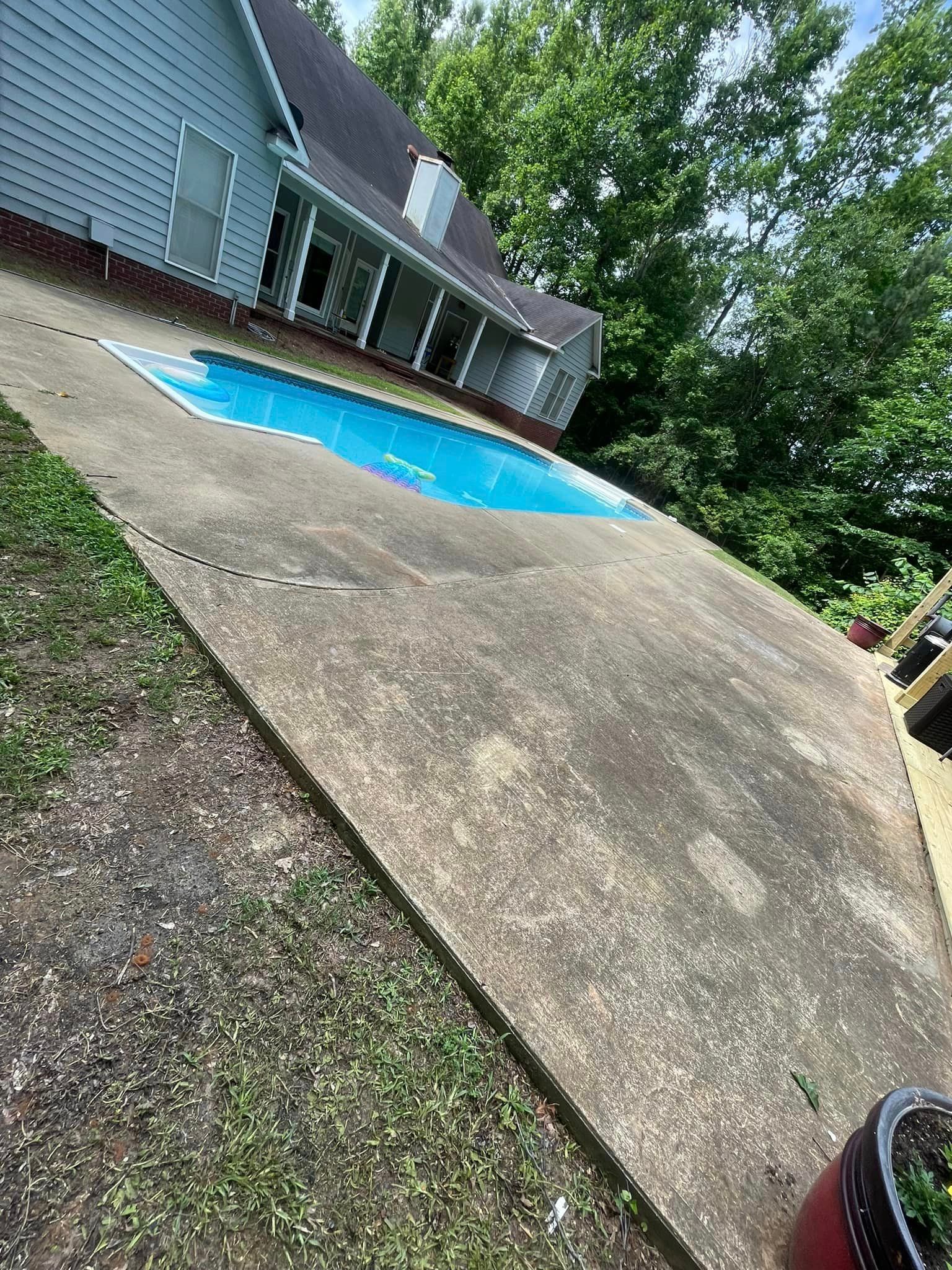 Concrete patio slopes down to a blue swimming pool beside a house. Lush green trees surround.