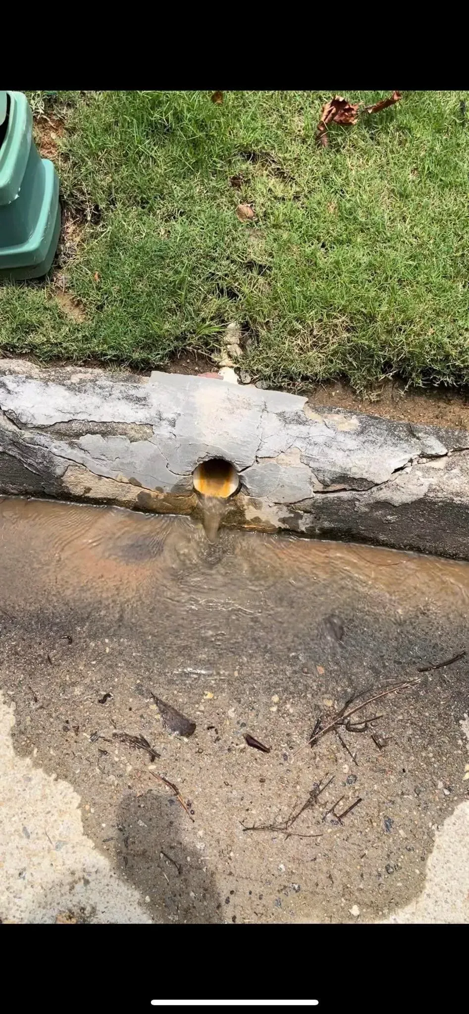 A drainpipe in concrete, discharging water onto asphalt near grass.