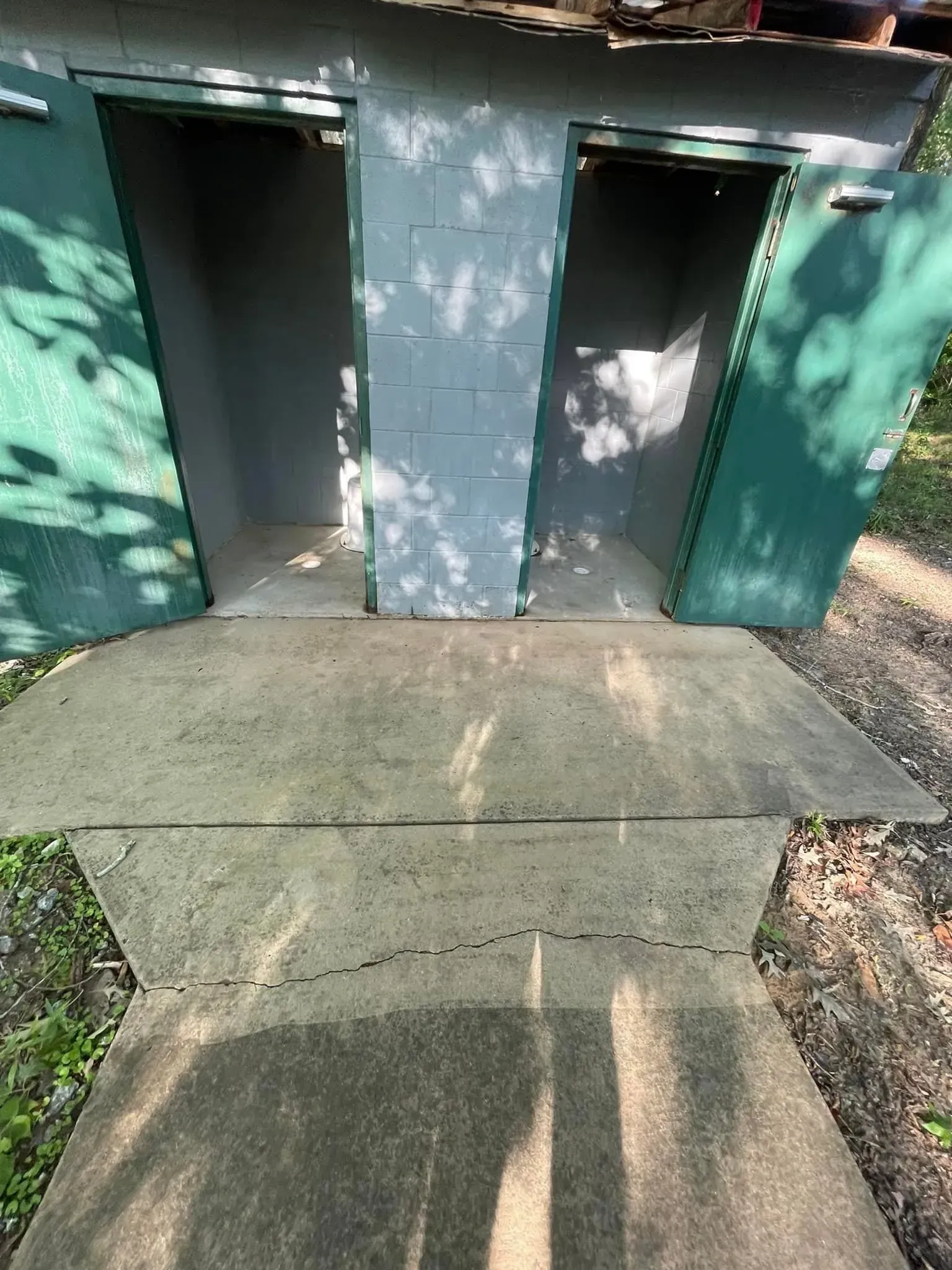Two open green door outhouses with a cracked concrete path.