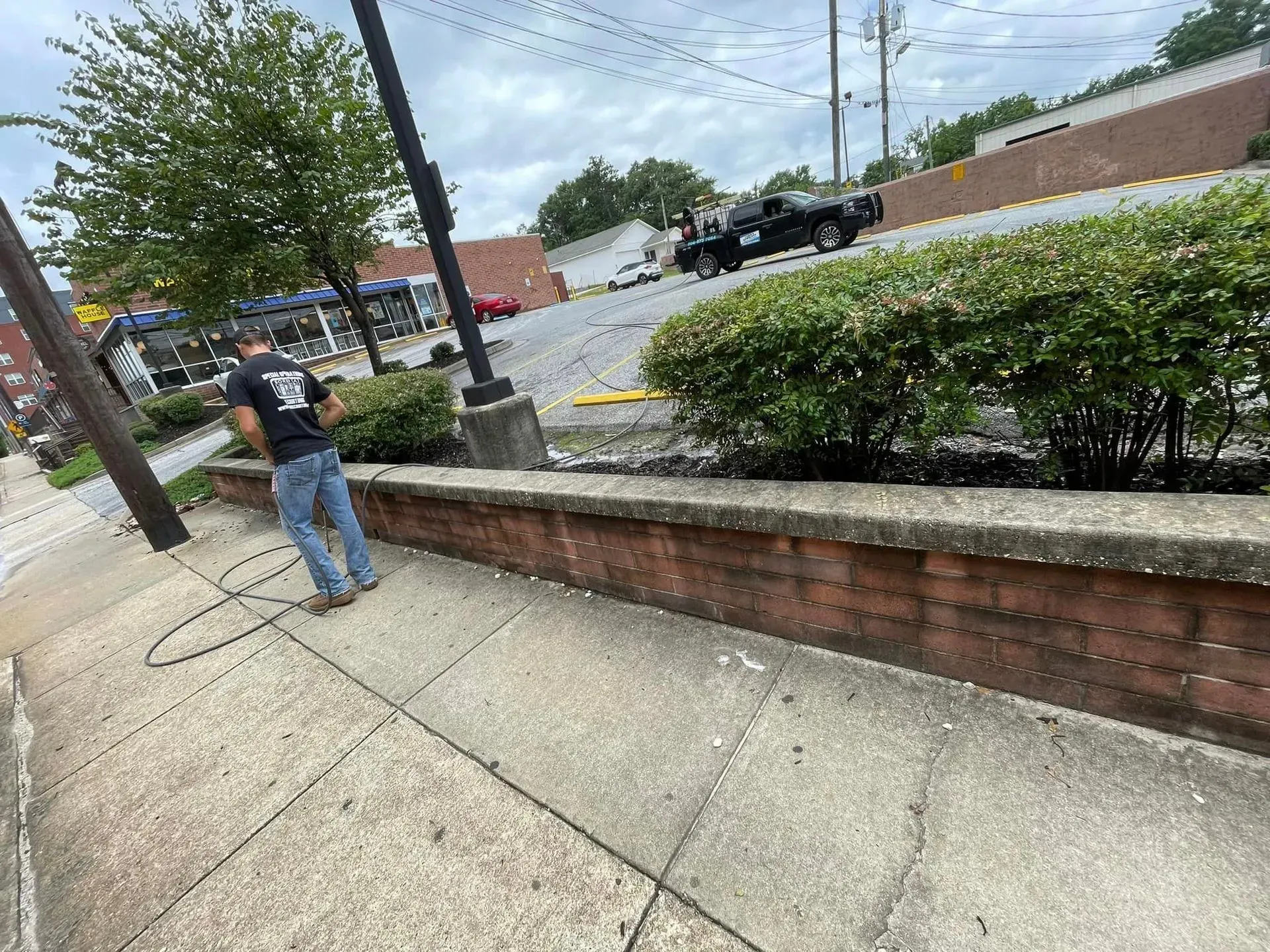 Person washing a brick wall with a hose on a sidewalk next to a raised garden bed with bushes.