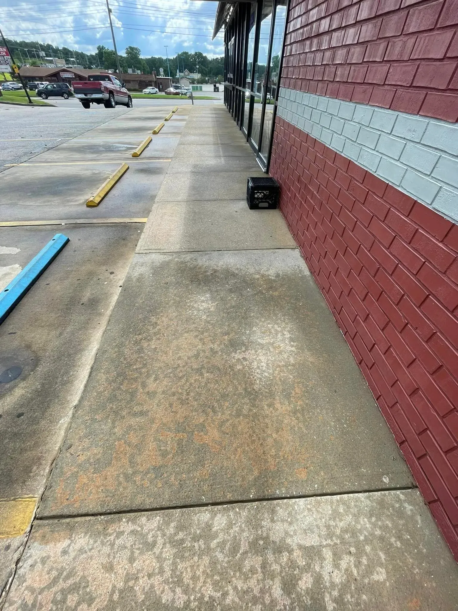 Sidewalk next to a brick building. Parking lot with parked cars and a blue parking block visible in the distance.