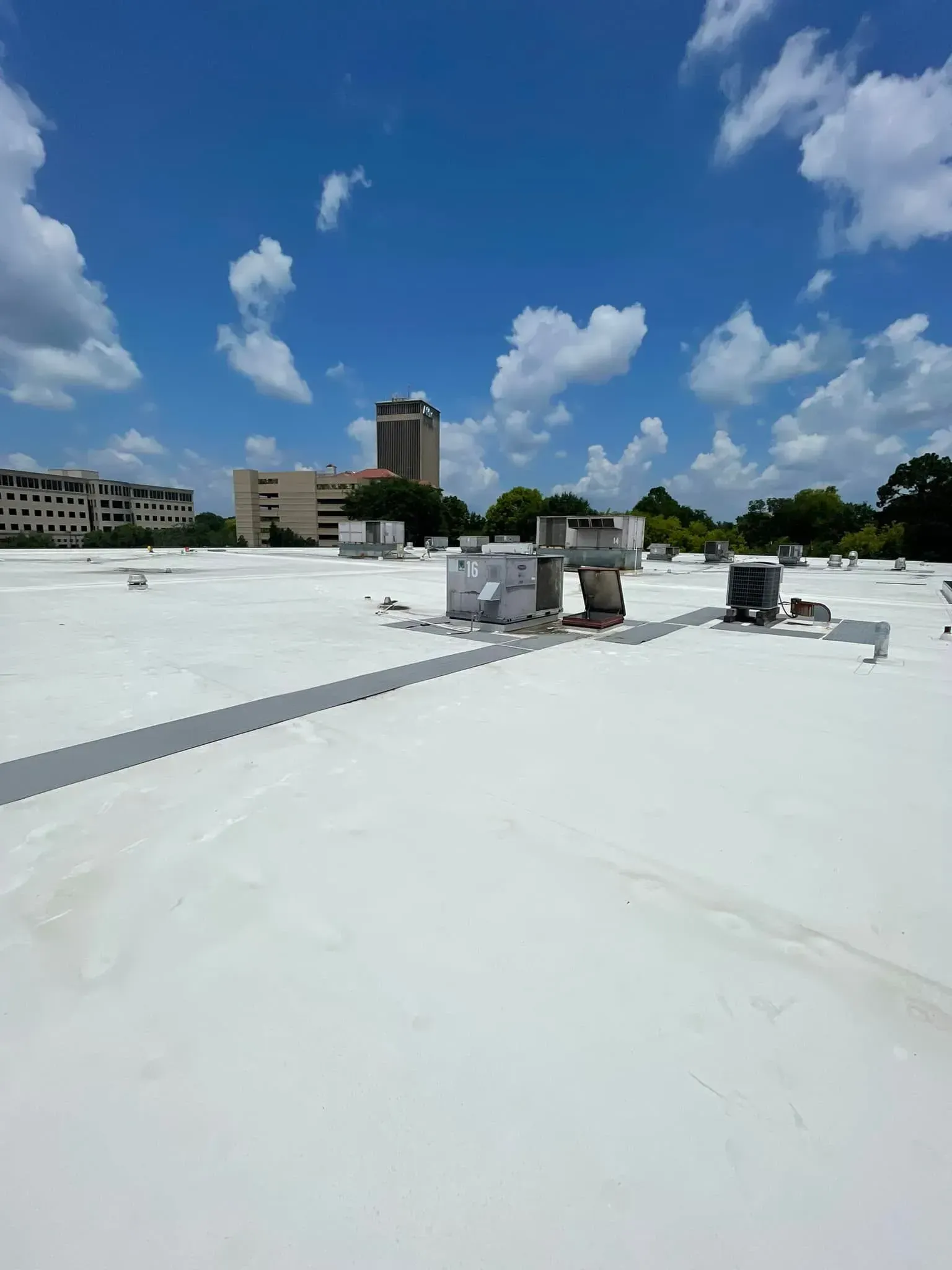 White flat roof with vents and city buildings under a bright blue sky with puffy clouds.