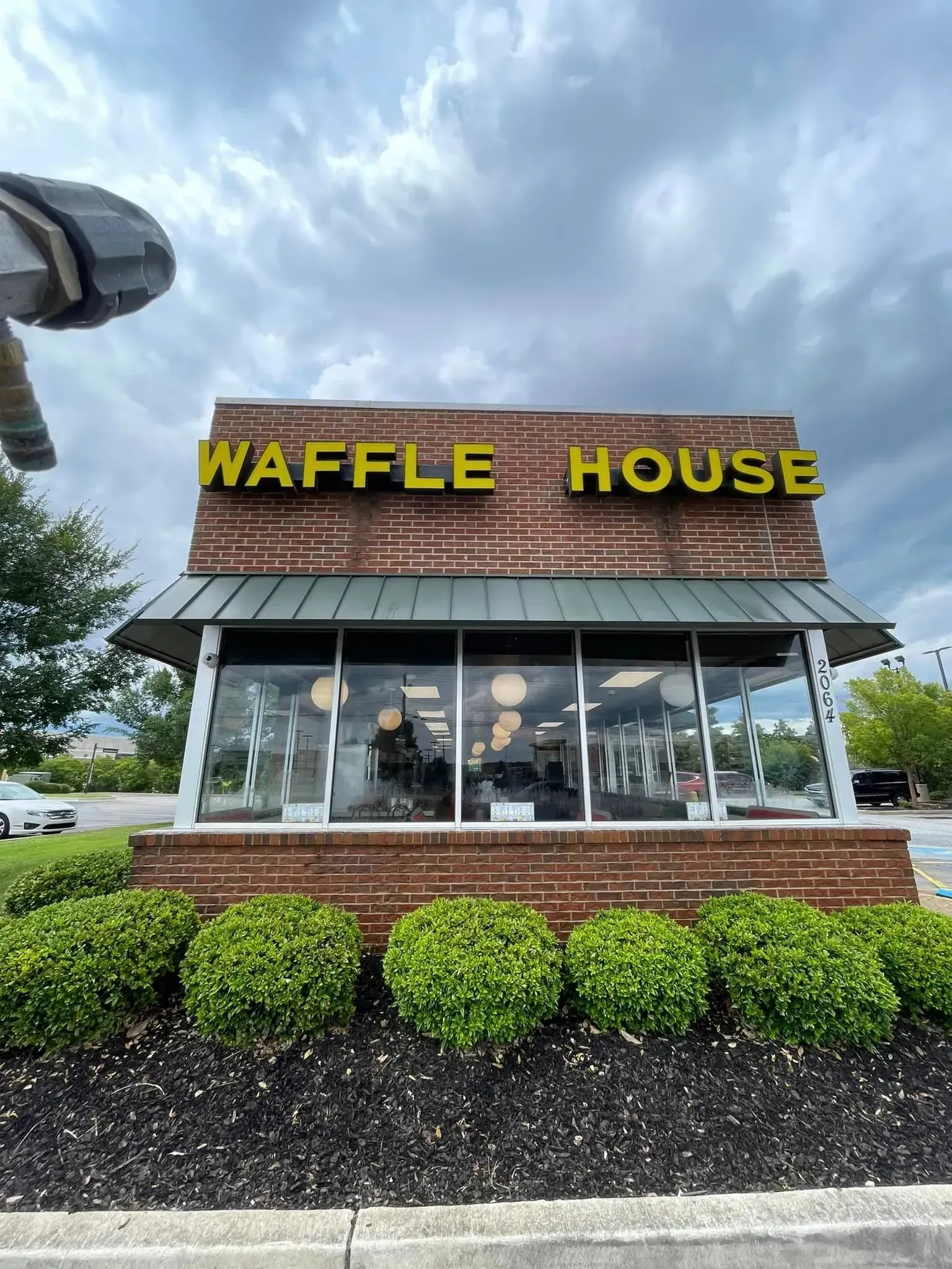 Waffle House restaurant with yellow sign, brick building, and green bushes against a cloudy sky.