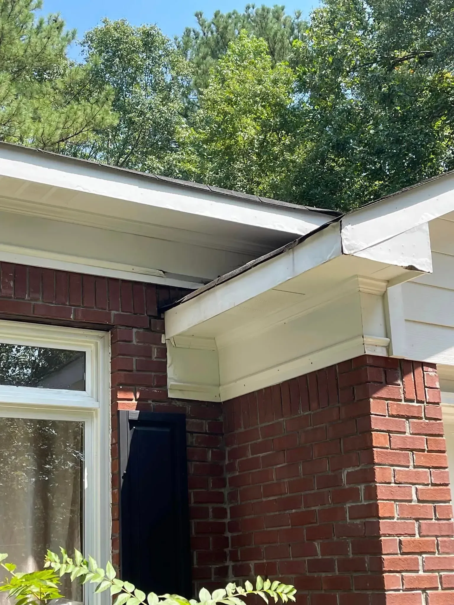 Corner of a brick building with white trim, roof, and soffits. Trees in the background, blue sky.