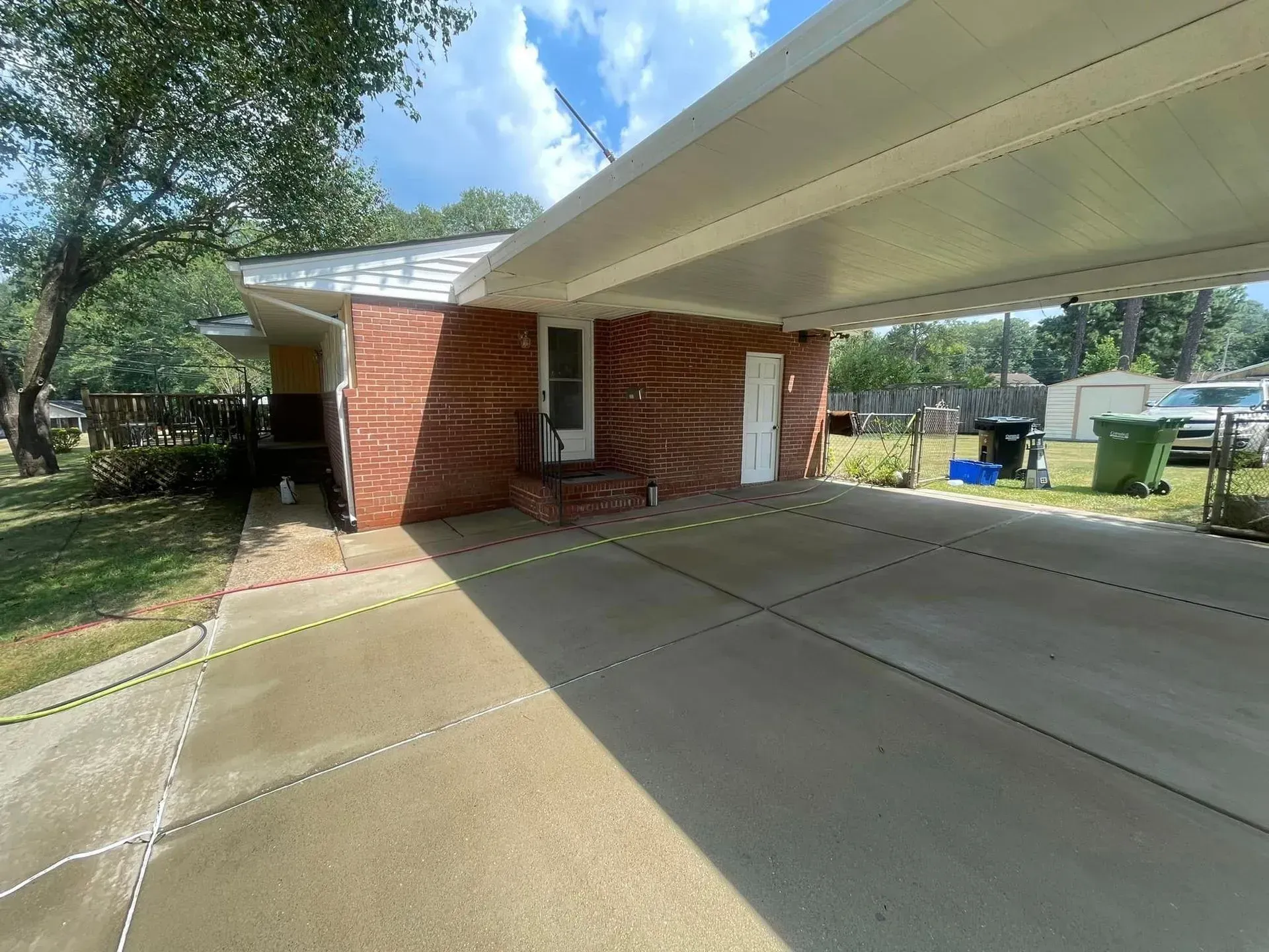 A red brick house with a carport on a sunny day. Concrete driveway and green grass visible.