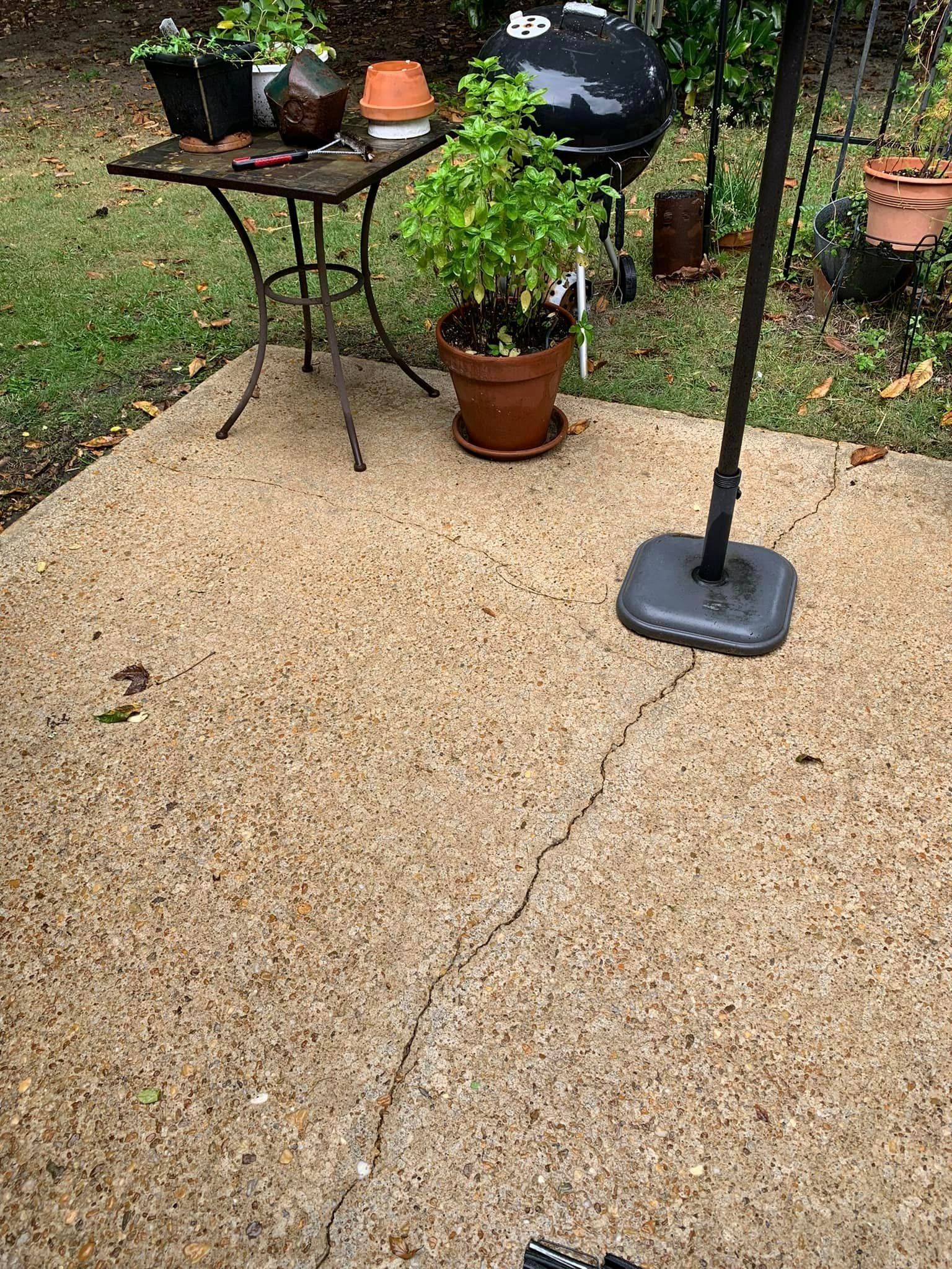 Concrete patio with a crack, small table, grill, and potted plants.