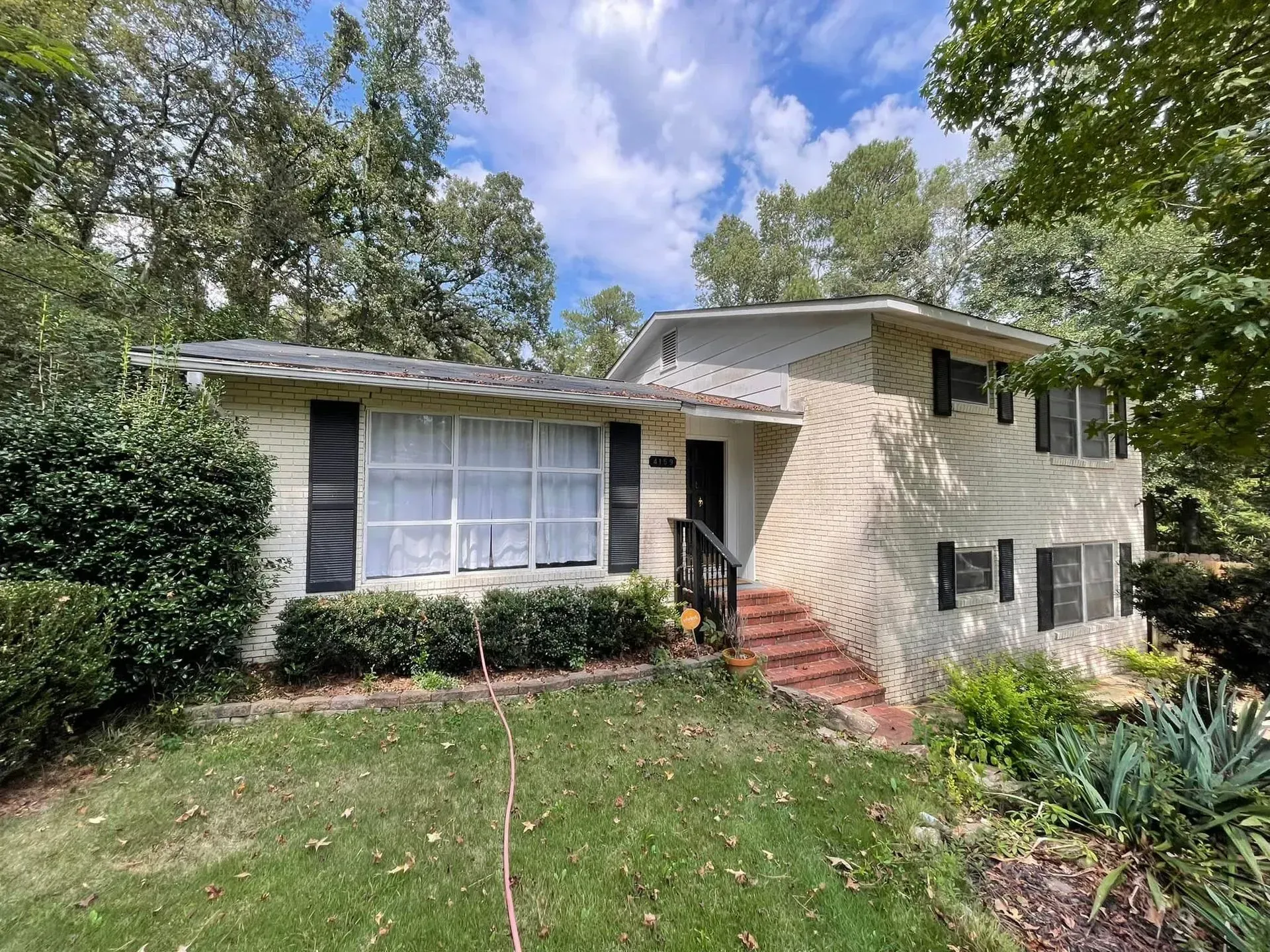 Two-story beige house with black shutters, brick steps, and a grassy yard. Trees surround the building.