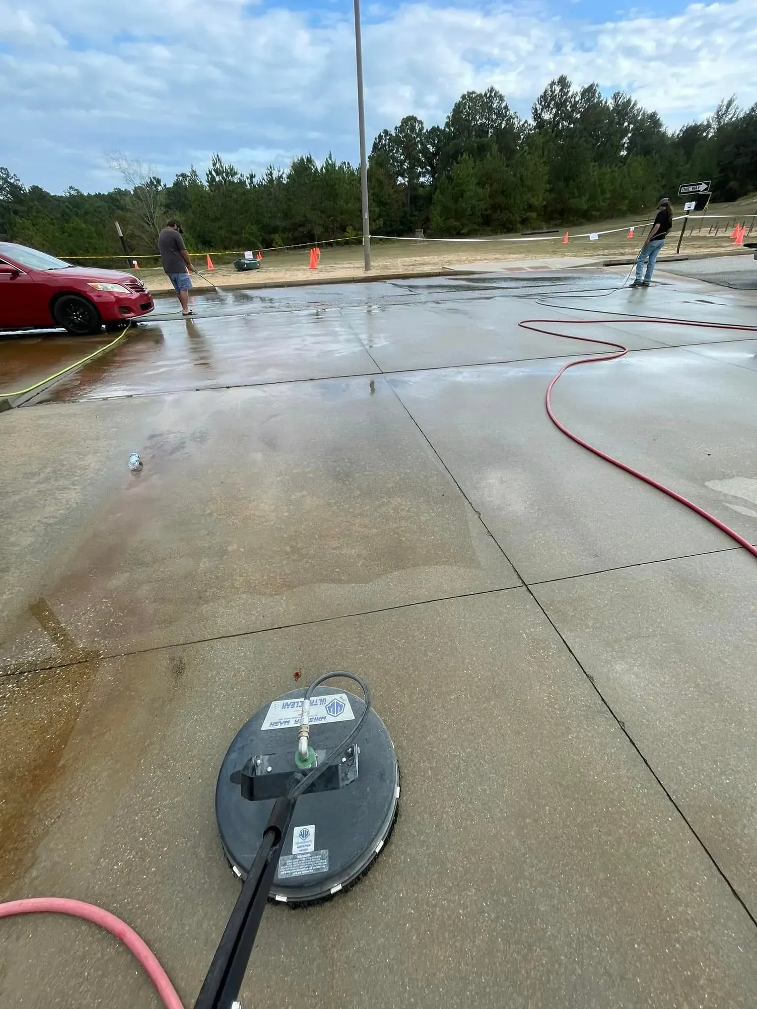 Pressure washer cleaning a wet concrete surface; two people and a red car in the background.