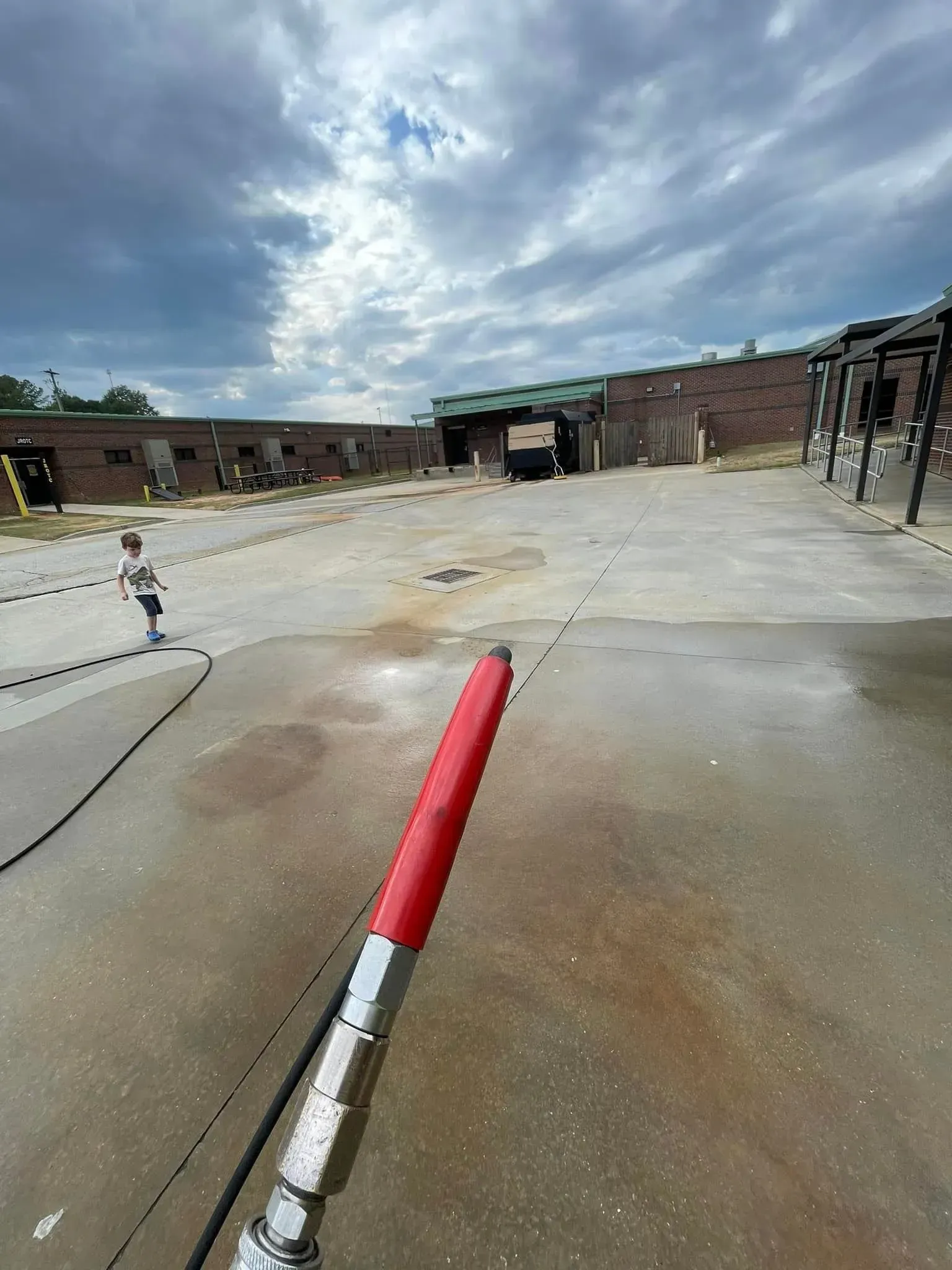 Person holding a red-tipped pressure washer nozzle, washing a concrete area in front of brick buildings under a cloudy sky.