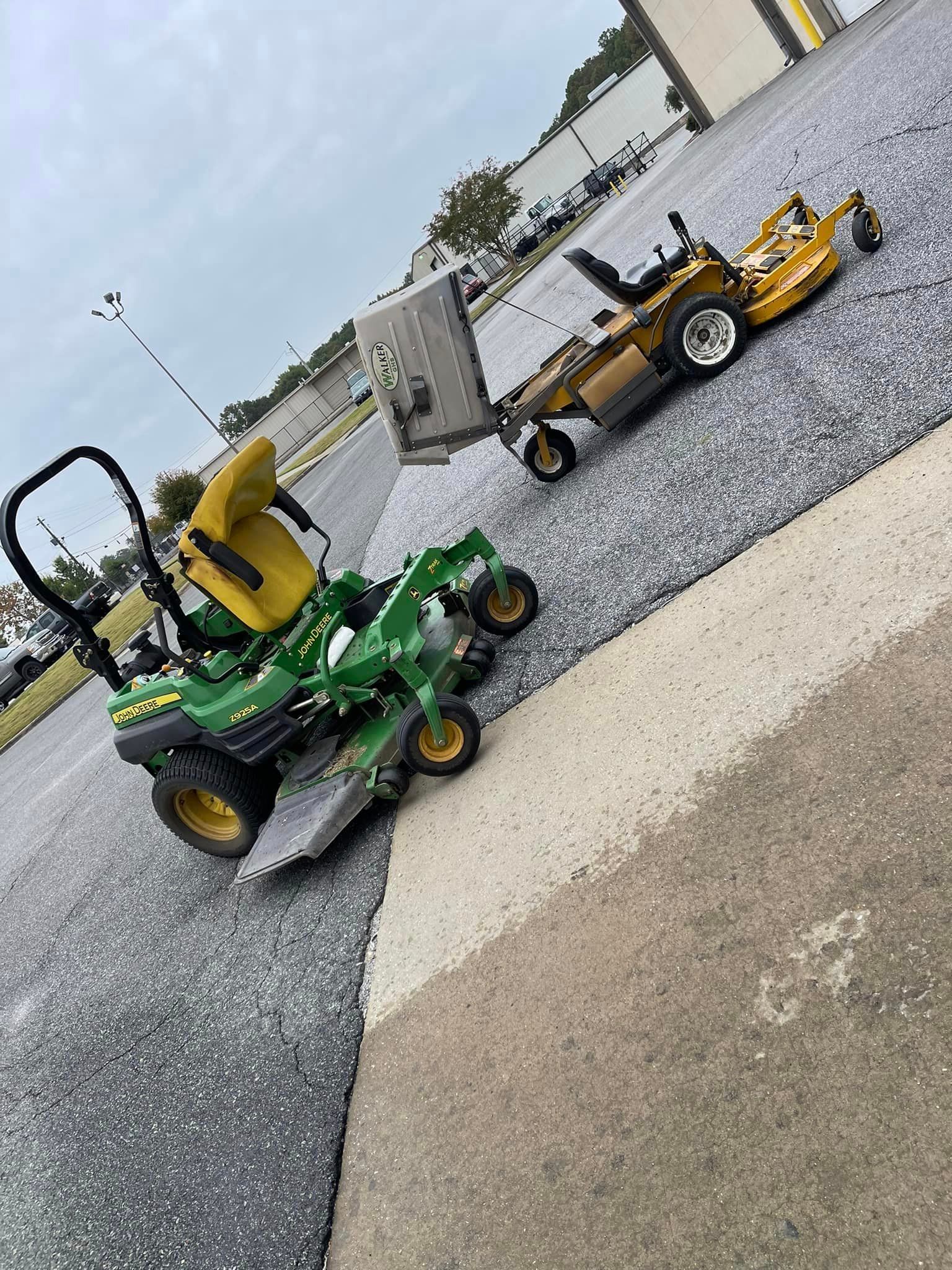 Two riding lawnmowers on a paved surface. One green and yellow, one gold and black. Overcast sky.