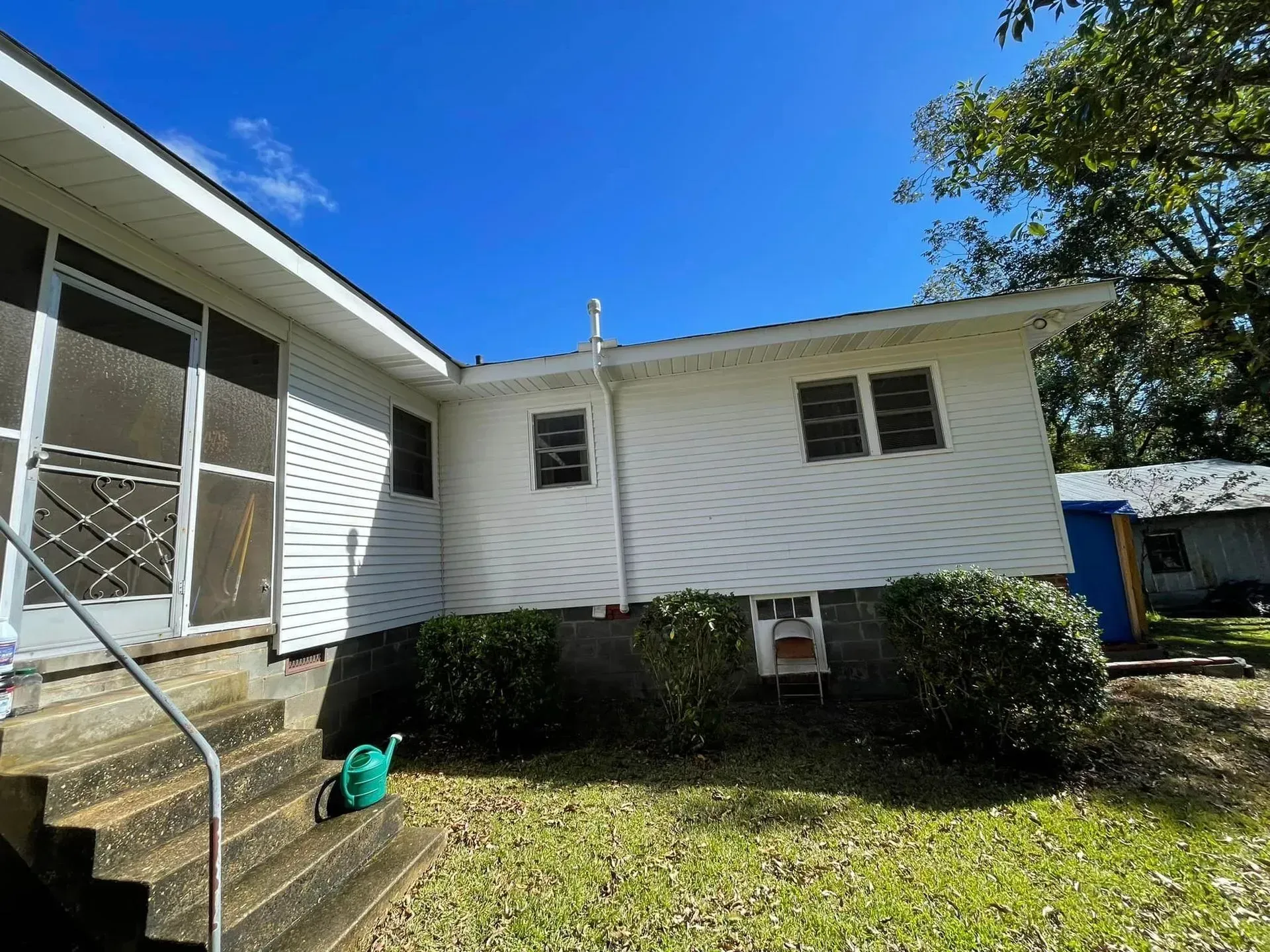 White house exterior with steps, screen porch, and small shrubs under the windows against a blue sky.
