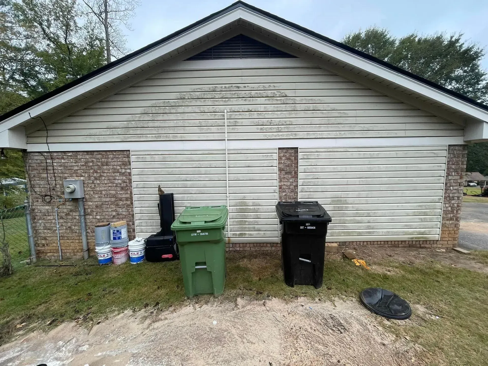 Garage with brick base, weathered siding, two trash bins, and paint supplies on the grass.