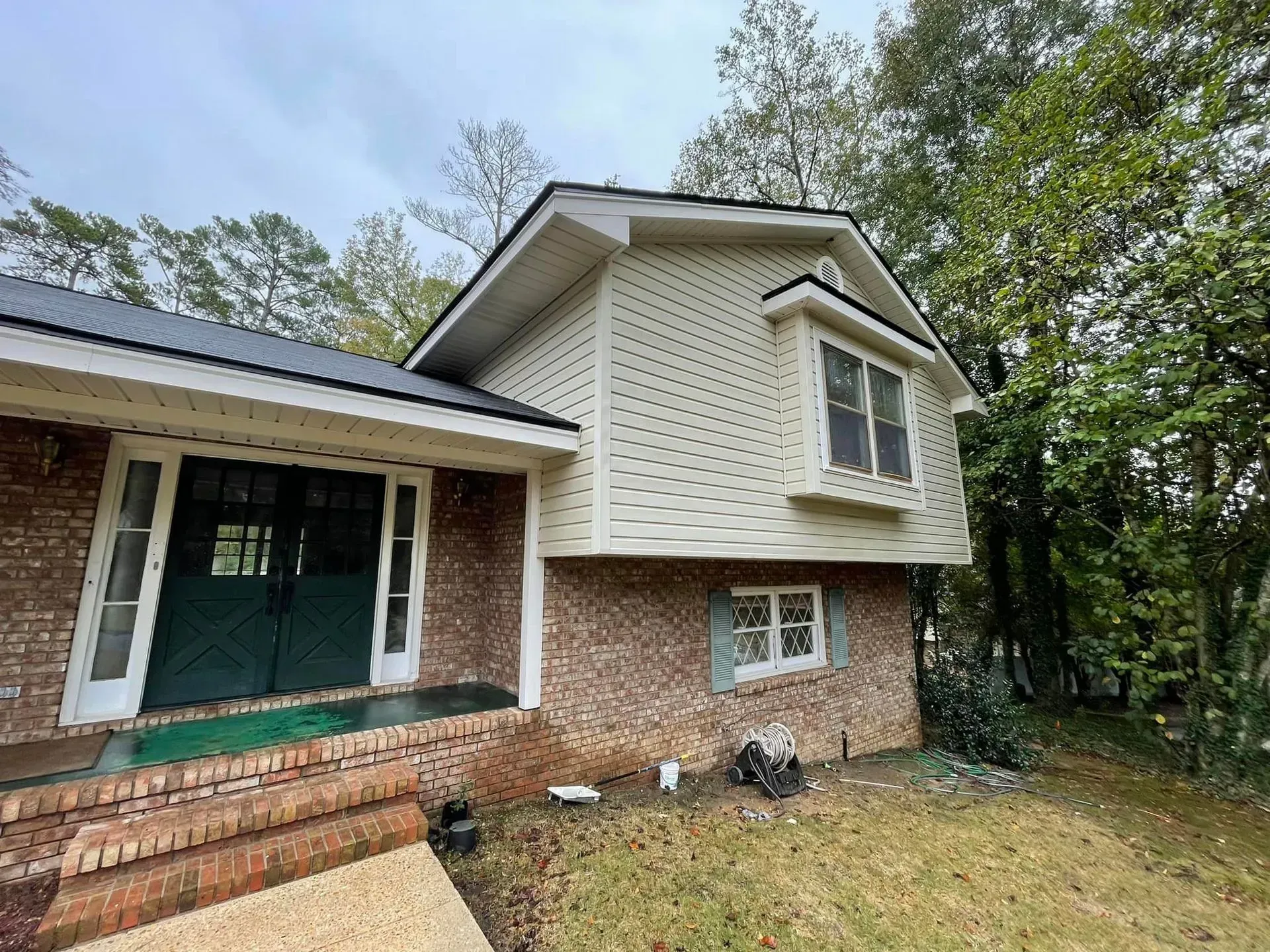 Brick and siding house with dark roof and green front doors, surrounded by trees on a cloudy day.