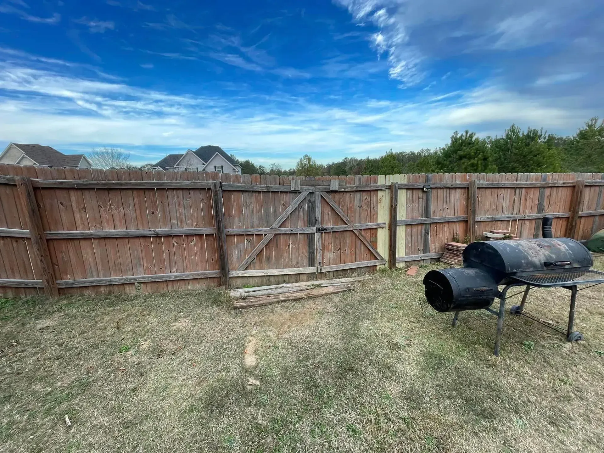 Backyard with a wooden fence, gate, smoker, and blue sky with clouds.