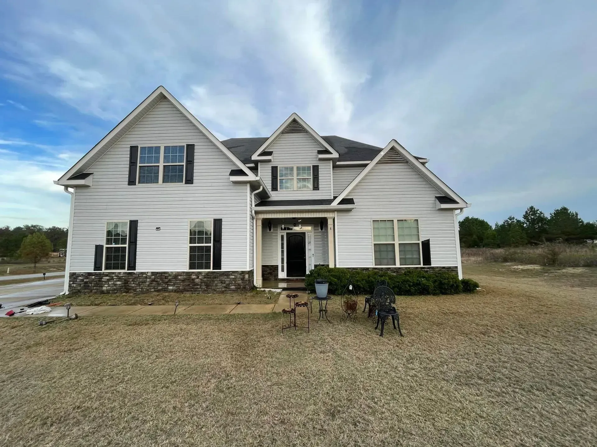 White two-story house with black shutters, brown trim, and a stone base, under a cloudy sky.