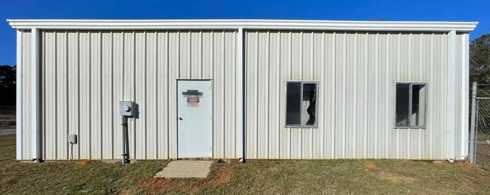 A white metal building with a door, two windows, and utility box; blue sky background.