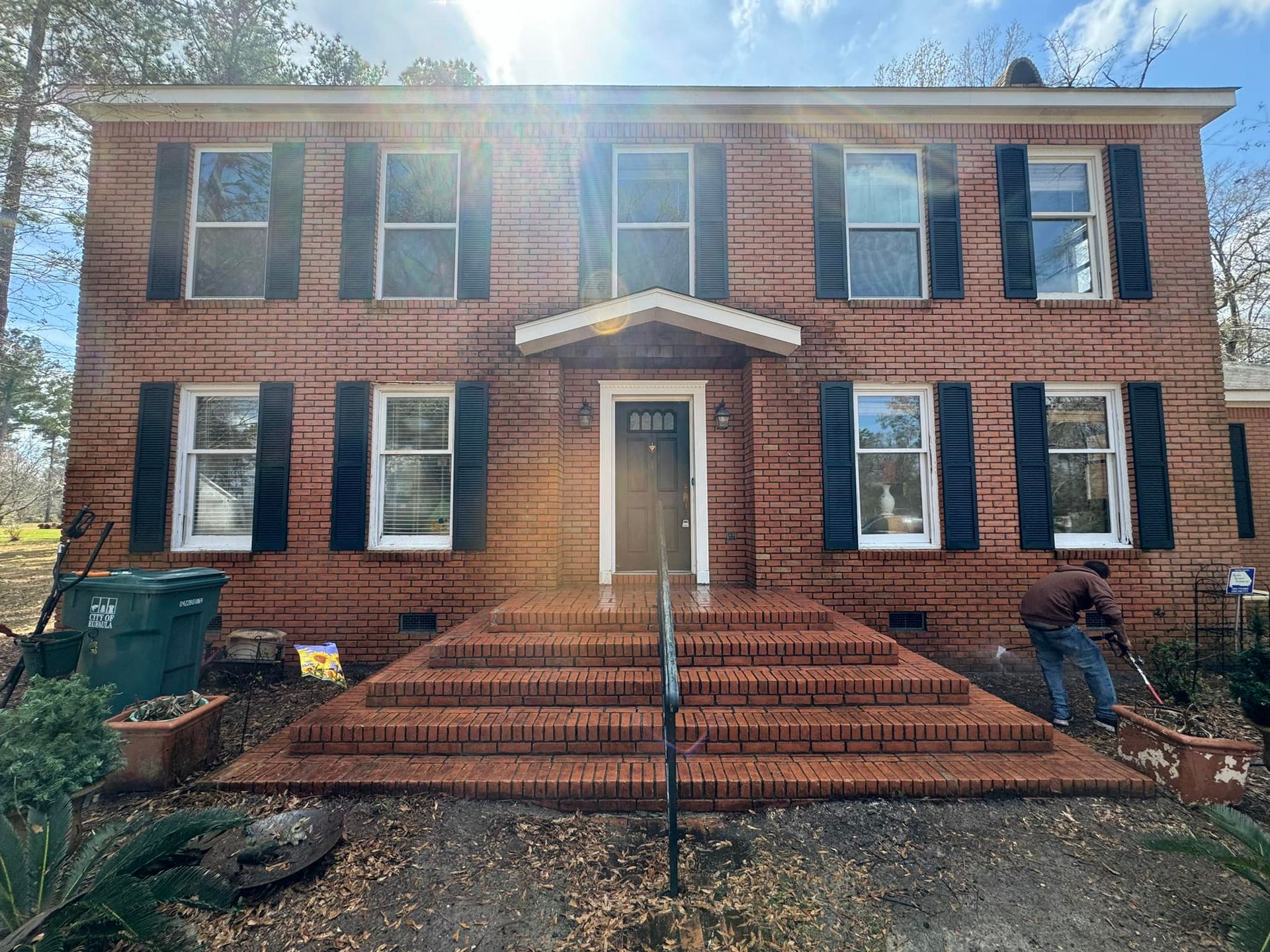 Red brick house with black shutters, steps, and person mowing.