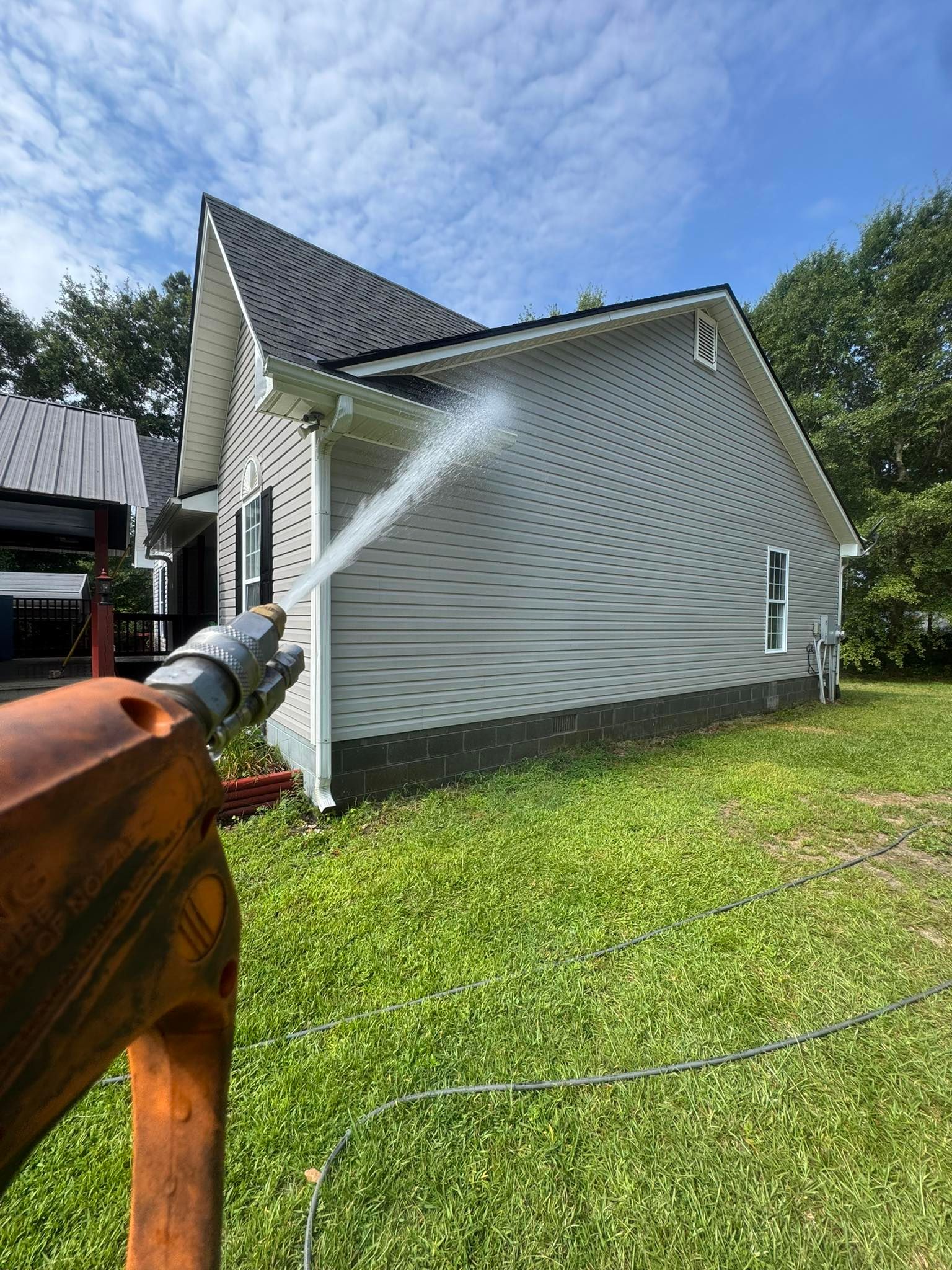 Pressure washing a gray vinyl-sided house with a black roof on a sunny day.