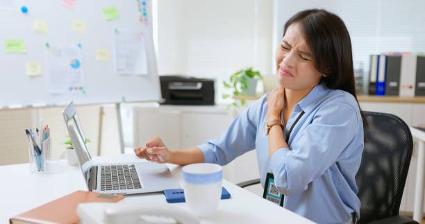 Woman in a blue shirt at a desk clutching her neck and grimacing in pain, likely due to poor posture while working on a laptop.