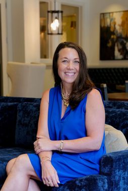 Woman in blue dress smiles, sitting on a blue couch; gold jewelry. Interior background.