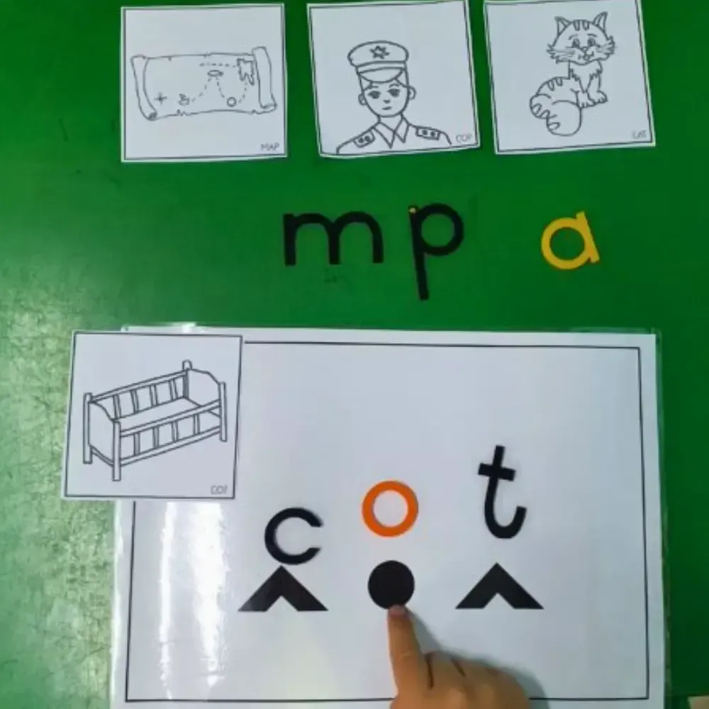 A child points at an orange dot while matching letters and images on a green surface.