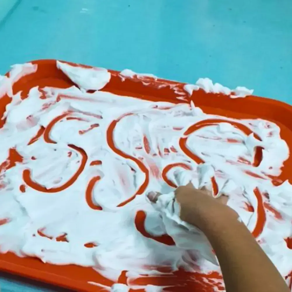 Child's hand writing letters in white shaving cream on an orange tray, against a blue background.