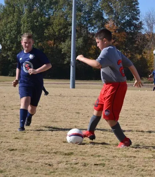 Two soccer players on a field, one in gray and red dribbling the ball, another in navy approaching.