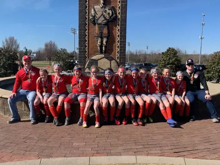 Group in red uniforms seated on a bench in a plaza, posing in front of a large statue.