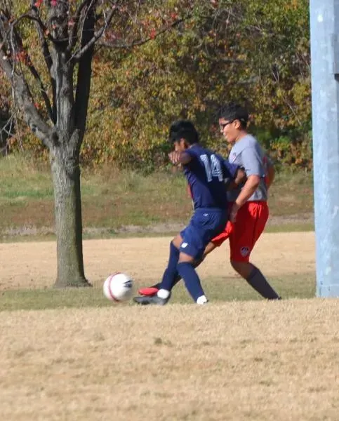 Two soccer players contest a ball on a grassy field, with autumn trees in the background.
