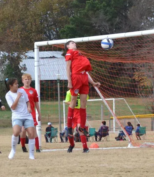 Soccer players jump for a header near the goal as the ball arcs toward the crossbar.