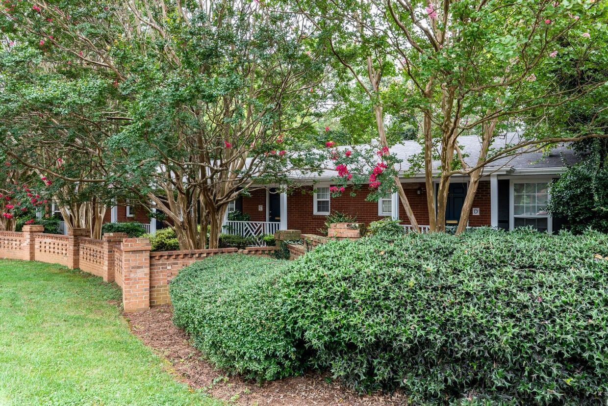 Low brick wall and green shrubs in front of a brick house with trees in the yard.