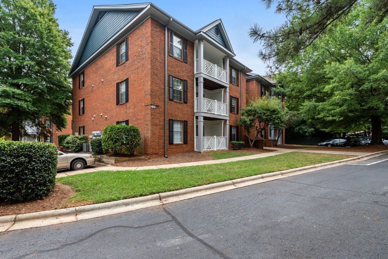 Brick apartment building with balconies, surrounded by trees and grass, on a sunny day.