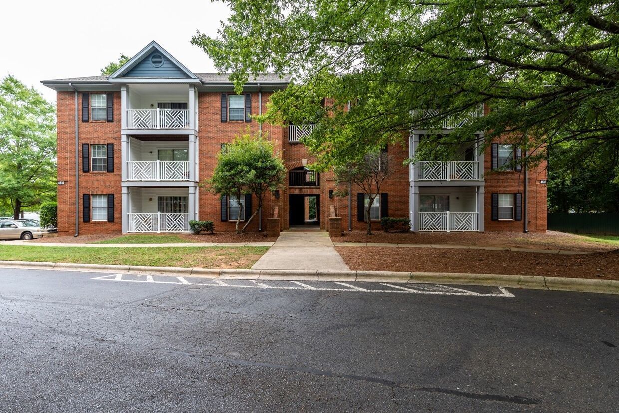 Apartment building with brick facade, balconies, and walkway. Trees and overcast sky in the background.