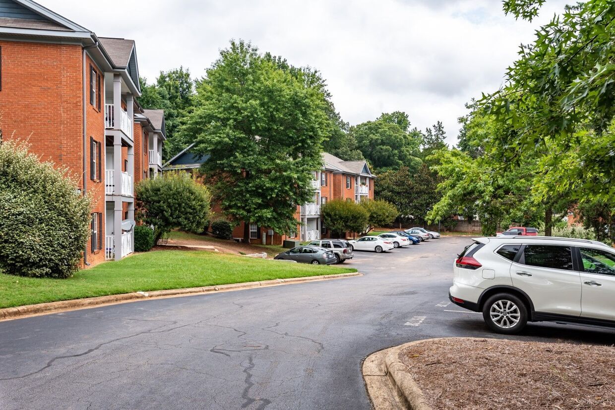 Apartment complex with brick buildings, parking lot with cars, and trees under a cloudy sky.