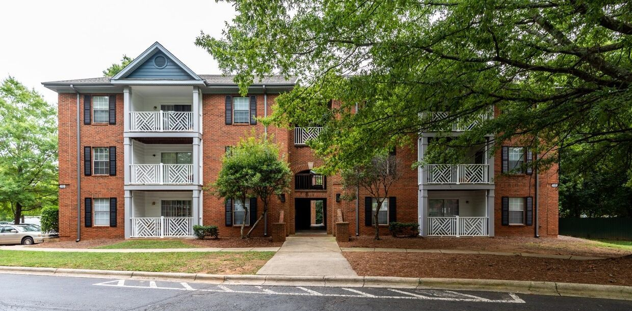 Brick apartment building with balconies, surrounded by trees and grass, on a sunny day.