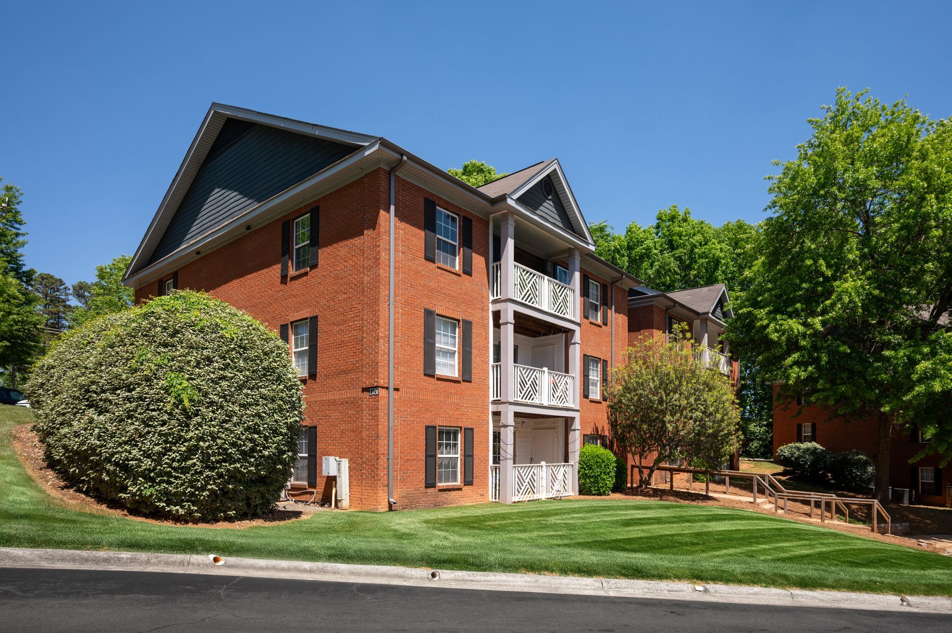 Brick apartment building with balconies, surrounded by trees and grass, on a sunny day.