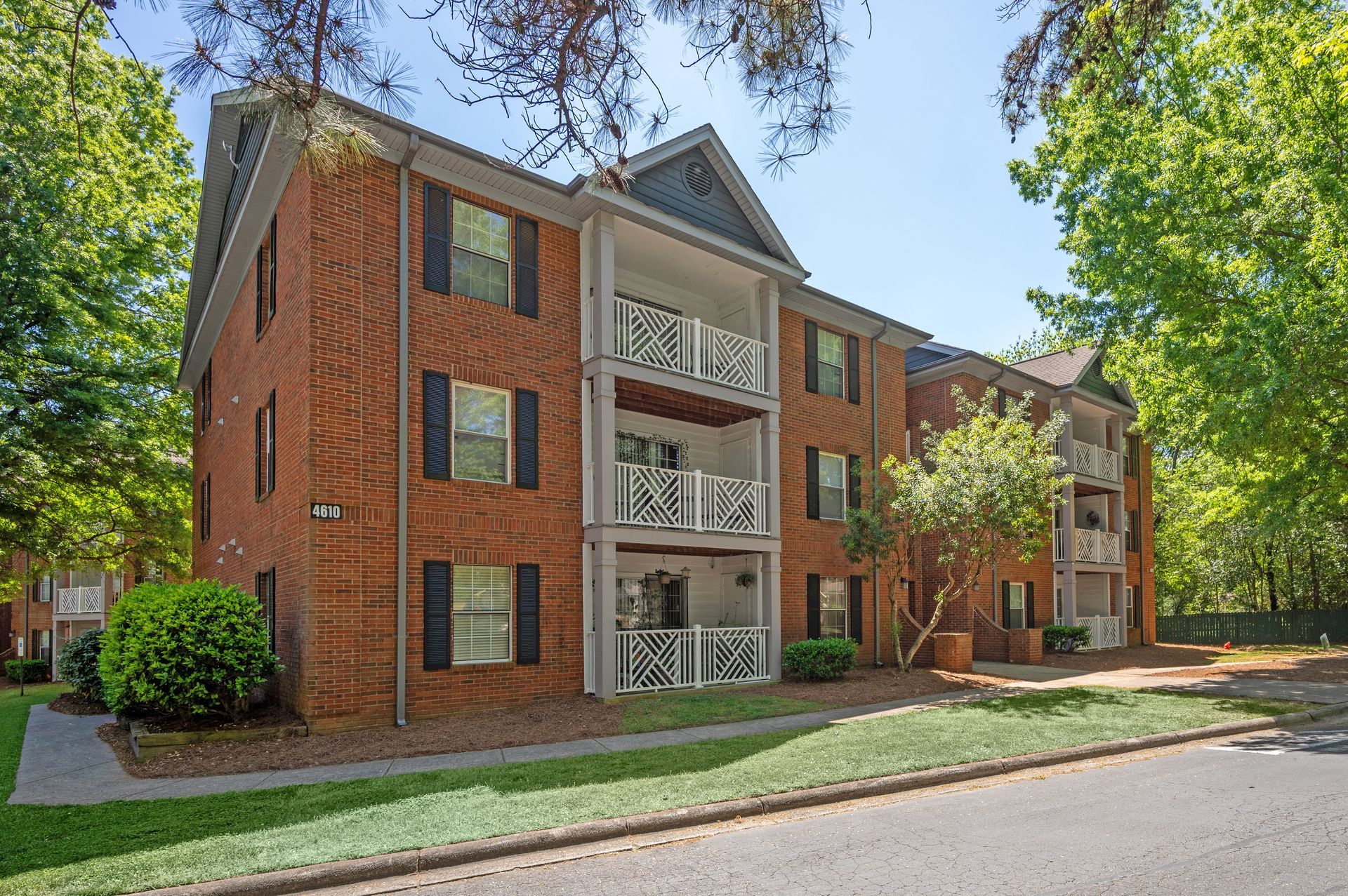 Brick apartment building with balconies, surrounded by trees and grass, on a sunny day.