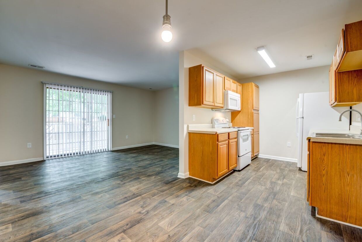 Kitchen with wood cabinets, white appliances, and gray flooring, open to a living area with sliding glass doors.