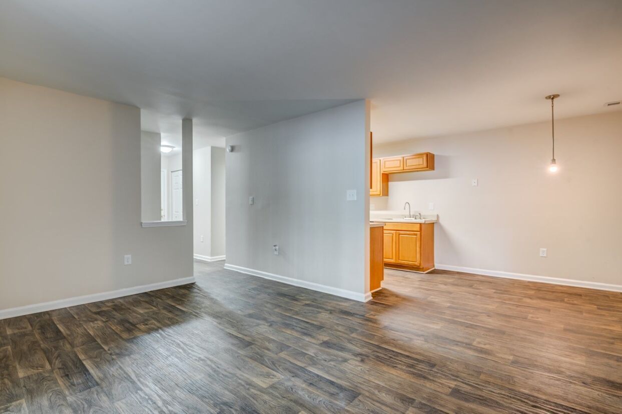 Empty apartment interior with wood flooring, light gray walls, and a kitchen visible in the background.