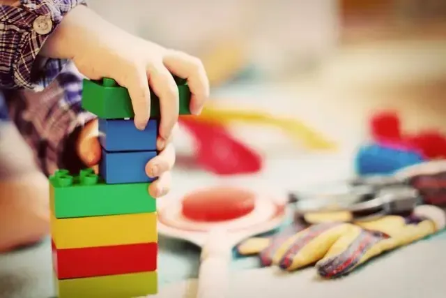 Child's hand stacking colorful toy blocks. Playroomin family assessment centre setting with other toys blurred in background.