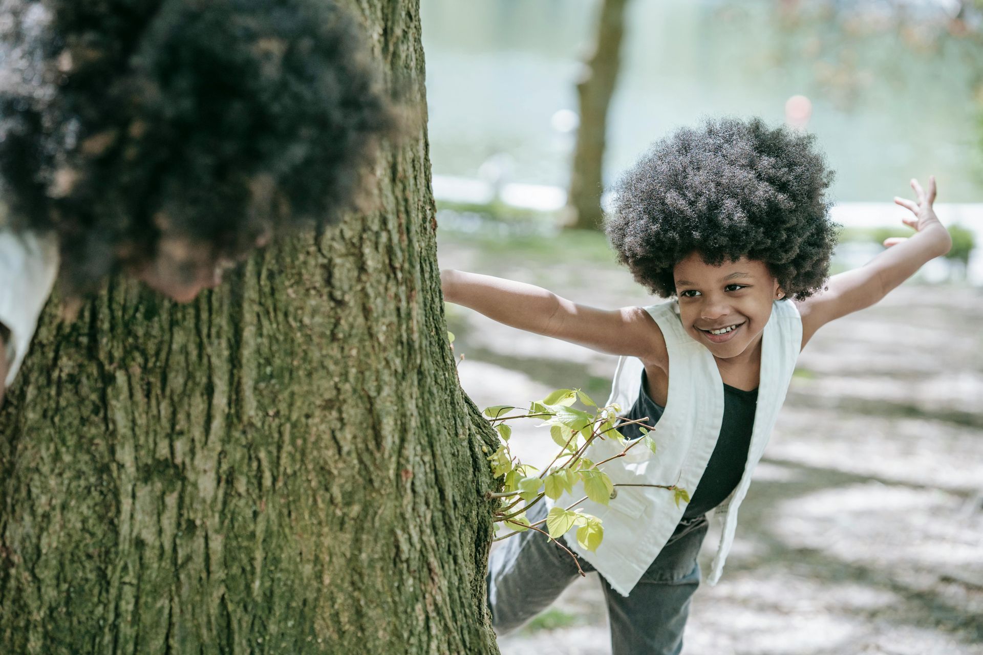 Child with large afro, smiling, arms outstretched, peeking around a tree trunk outdoors.